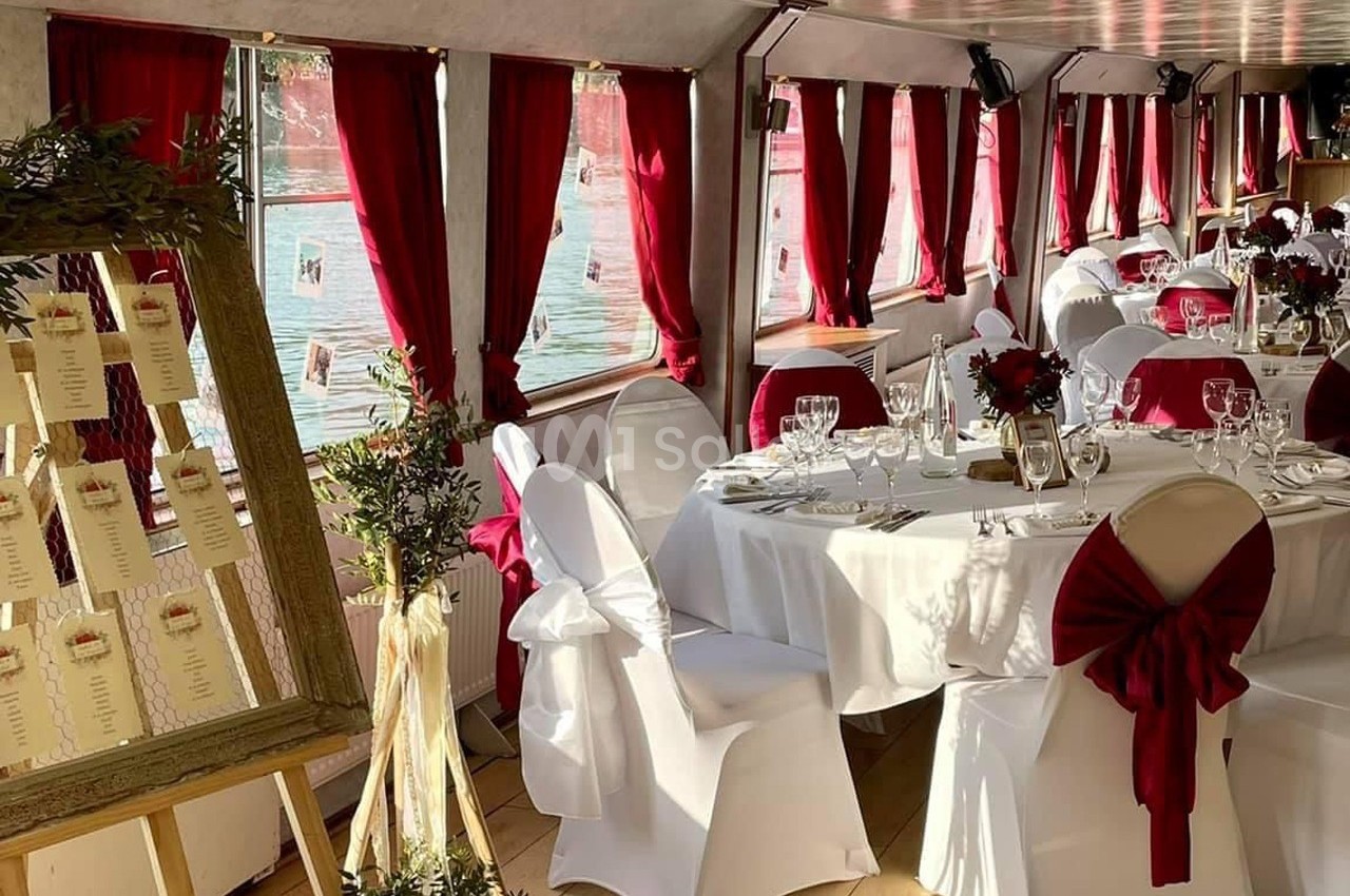 Salle de réception décorée avec des nappes blanches, des rubans rouges et un plan de table sur chevalet.