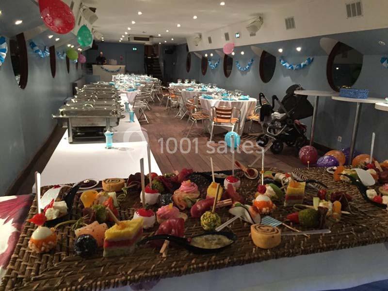 Buffet de nourriture variée en premier plan avec une salle décorée et des tables dressées en arrière-plan.