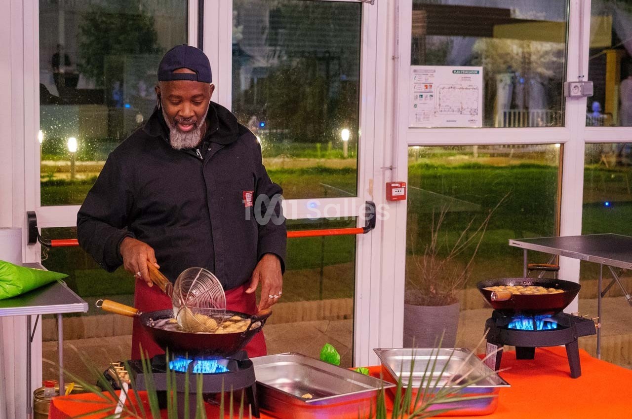 Un cuisinier souriant prépare des plats frits dans des poêles sur des réchauds, dans un espace lumineux et vitré.
