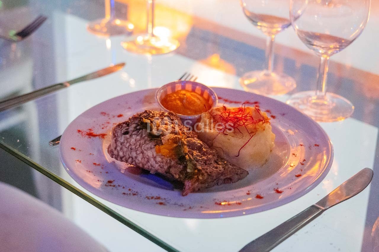 Assiette de poisson grillé avec purée, sauce et garniture, servie sur une table en verre avec couverts et verres à vin.