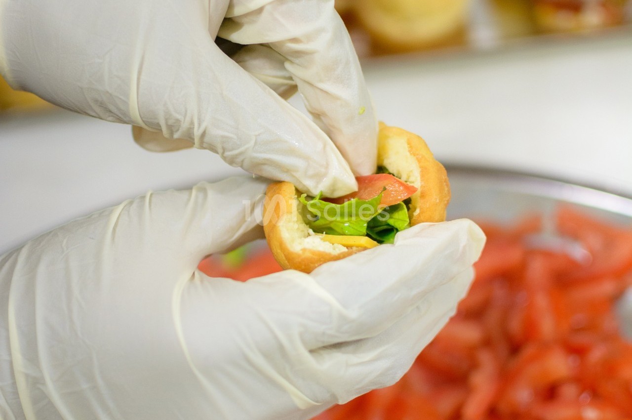 Une main gantée garnit un petit pain avec de la salade et une tranche de tomate, en cuisine.