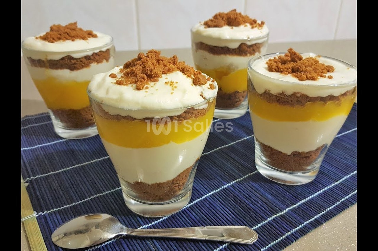 Verrines composées de couches de spéculoos, crème, et coulis de mangue, présentées sur une nappe bleue avec une cuillère.