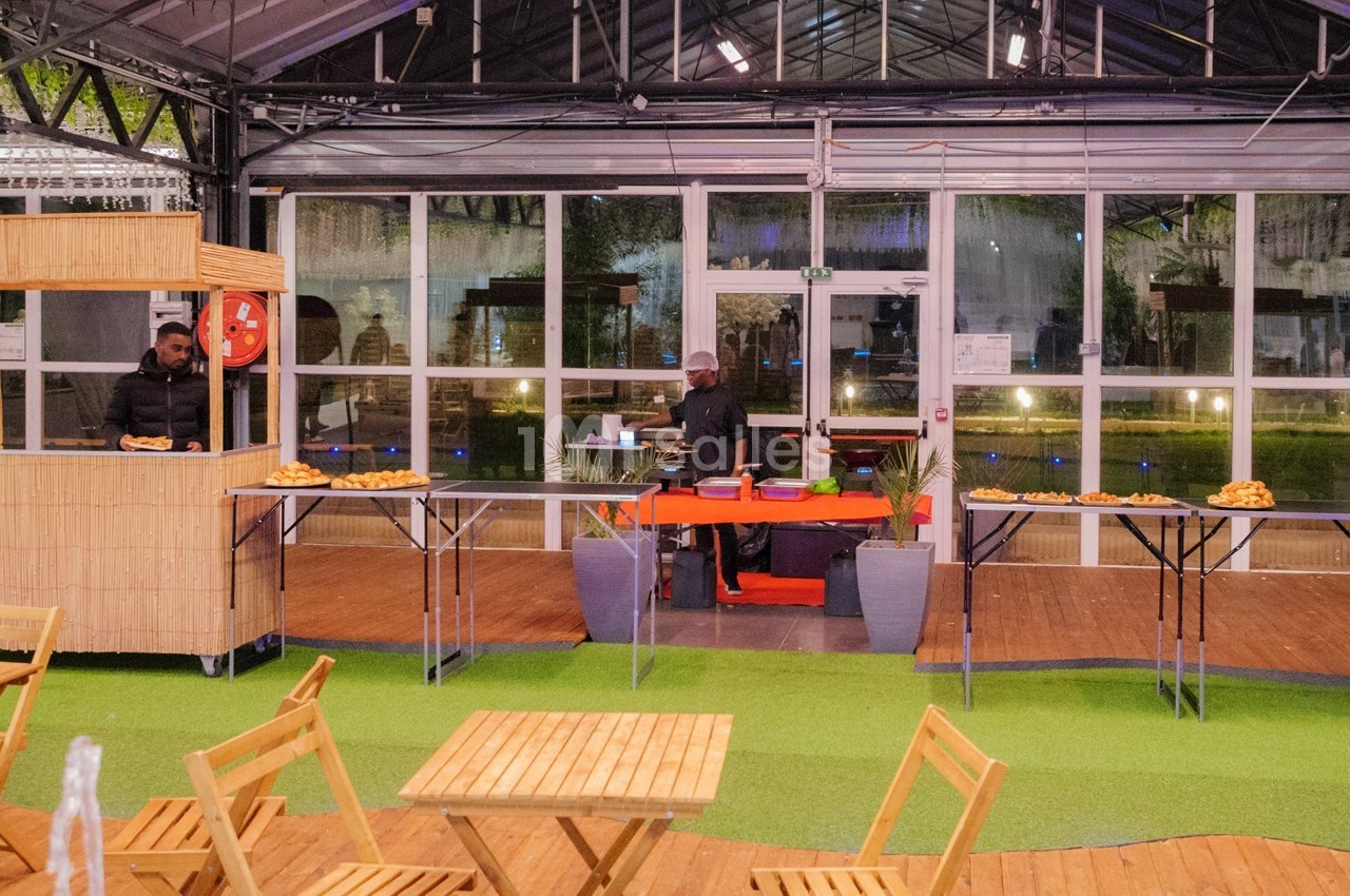 Un stand de restauration avec deux cuisiniers, des tables en bois et des plats disposés sur des tables dans un espace…