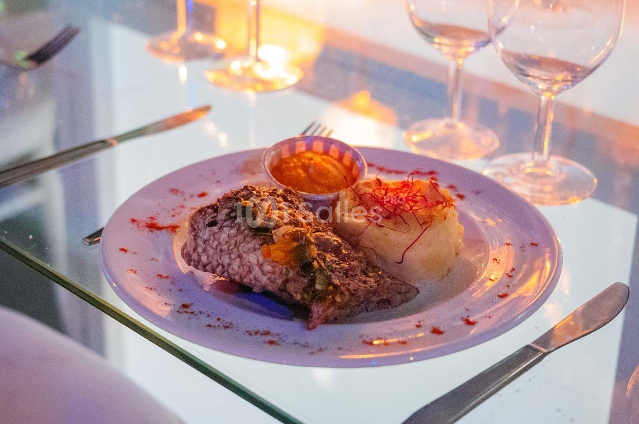 Assiette de poisson grillé avec purée, sauce et garniture, servie sur une table en verre avec couverts et verres à vin.