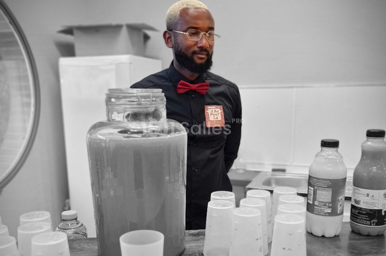 Un homme derrière un comptoir avec des boissons en bouteilles et un grand récipient de jus devant lui.
