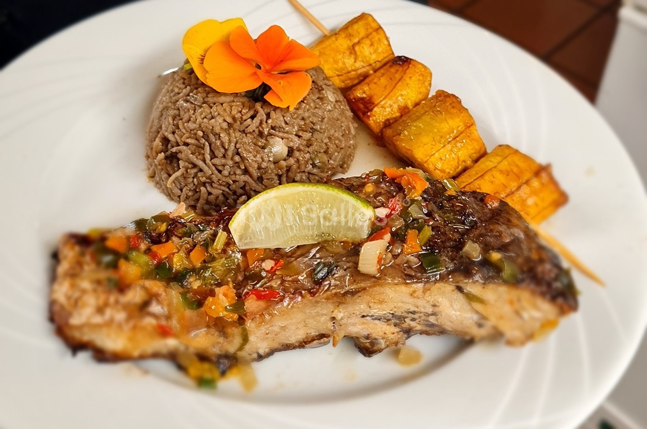 Assiette composée de poisson grillé avec sauce, riz aux haricots, brochettes de bananes plantains et une fleur décorative.