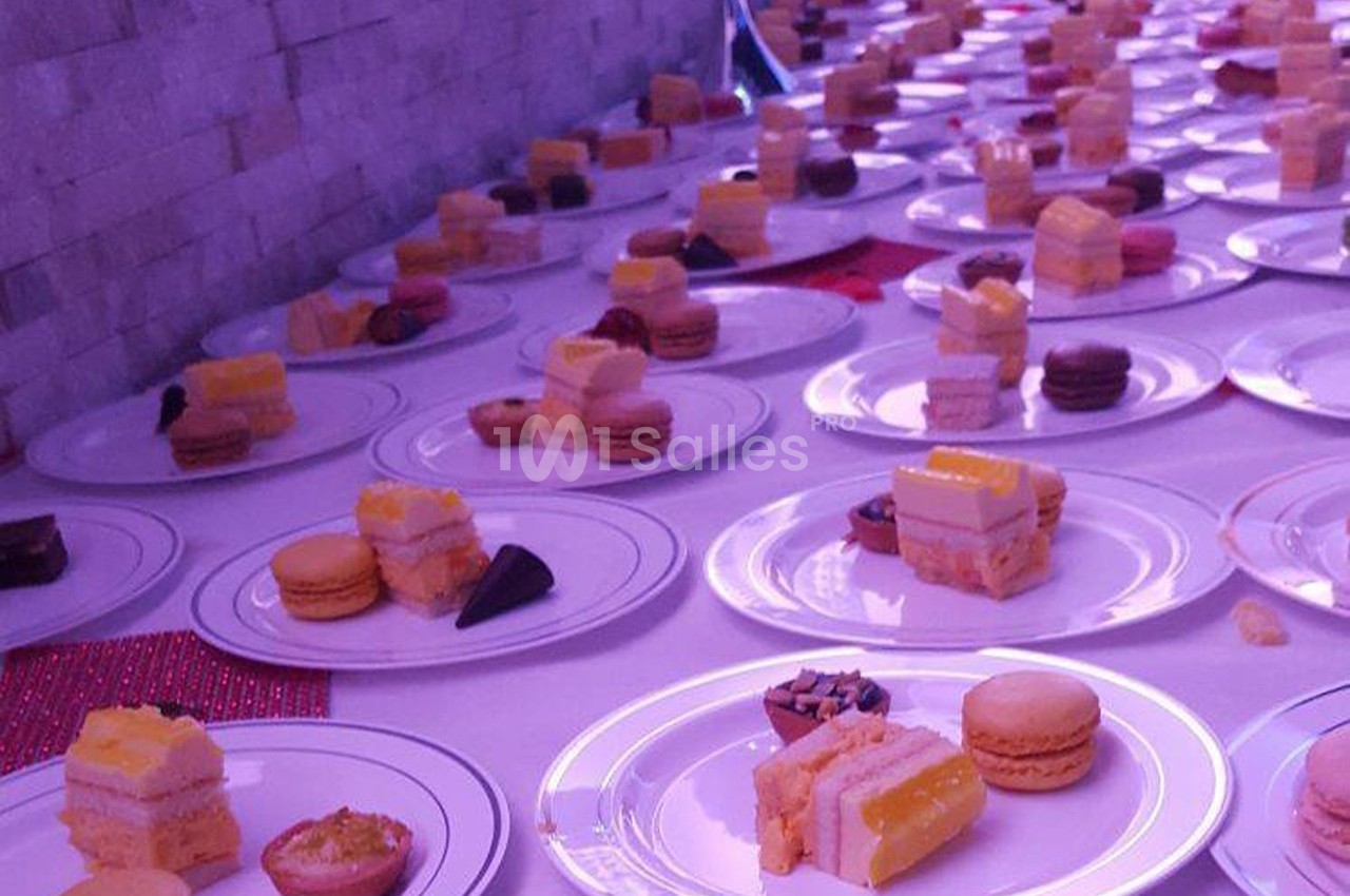 Assiettes alignées contenant des desserts variés, dont macarons, gâteaux et pâtisseries, sur une table éclairée en violet.