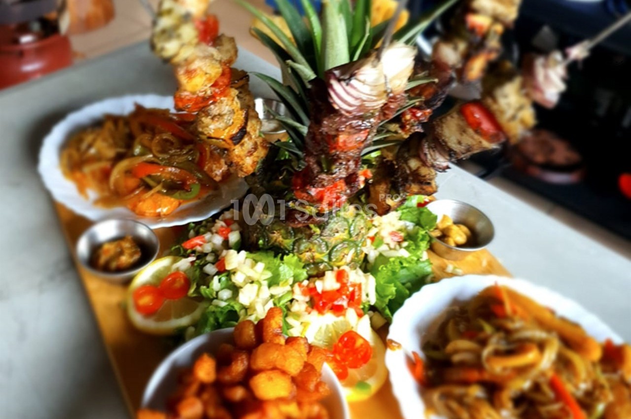 Assiette garnie de brochettes sur un ananas, accompagnée de légumes, sauces et bouchées frites.