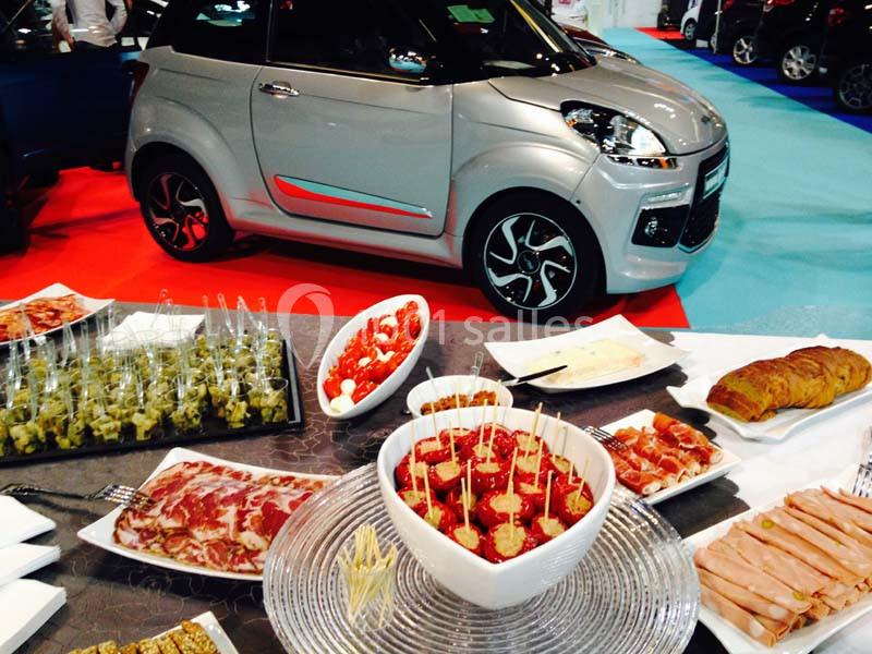 Buffet varié avec charcuterie, amuse-bouches et viennoiseries en premier plan, petite voiture grise en arrière-plan.