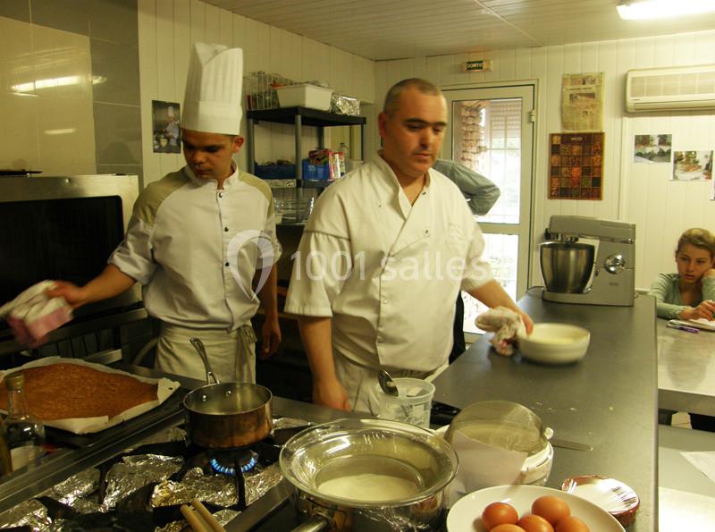 Deux cuisiniers en tenue blanche préparent des plats dans une cuisine professionnelle équipée.