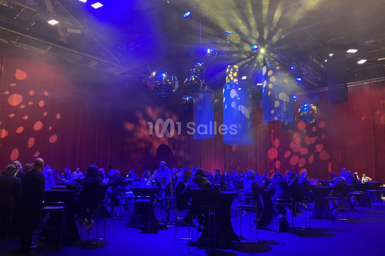 Salle de réception avec invités assis à des tables hautes, éclairée par des lumières colorées et des boules réfléchissantes.