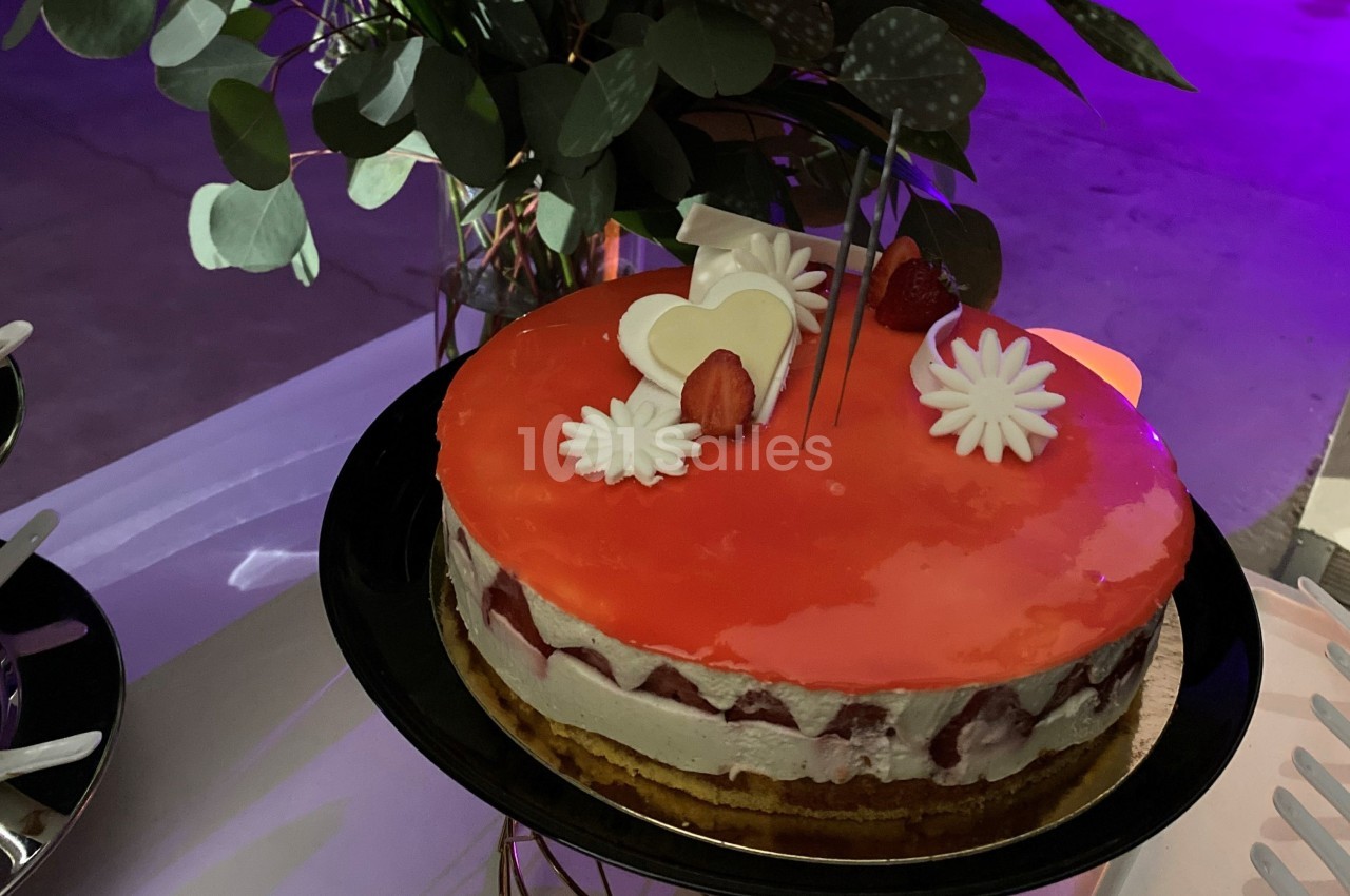 Gâteau avec glaçage rouge, décoré de fraises, fleurs en sucre et un cœur blanc, posé sur une table.
