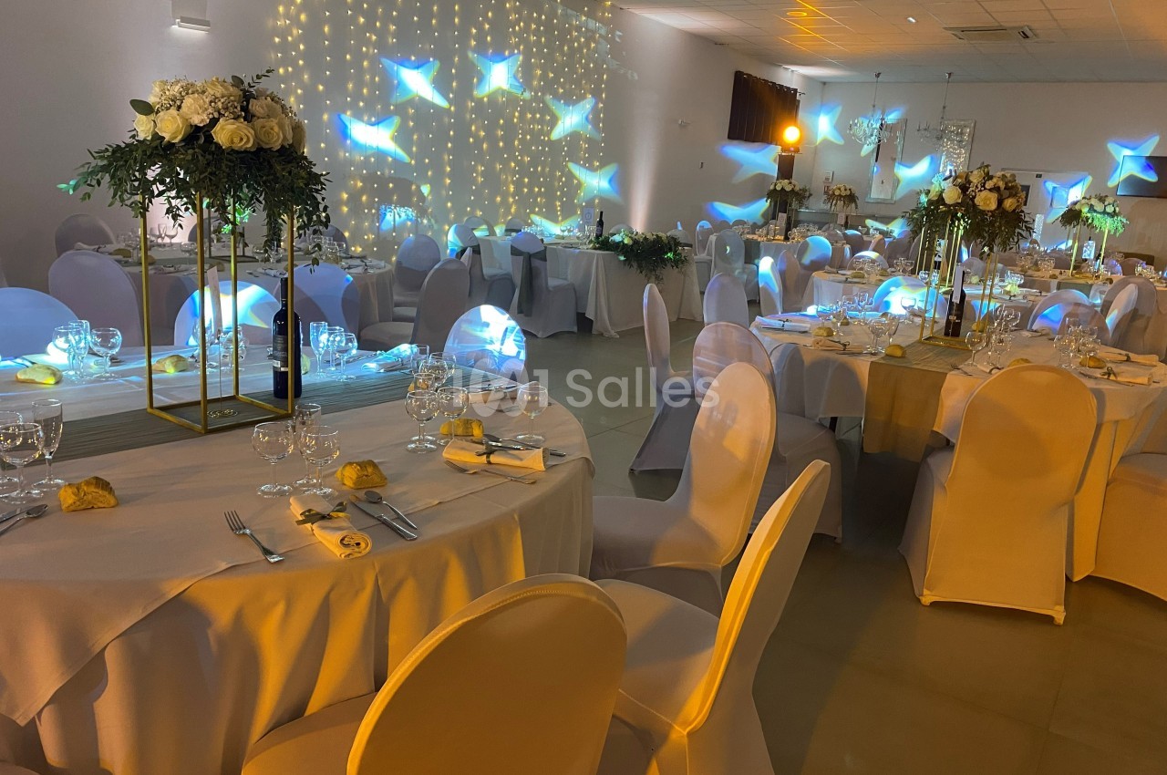 Salle de réception décorée pour un événement, avec tables dressées, chaises blanches et éclairage lumineux.