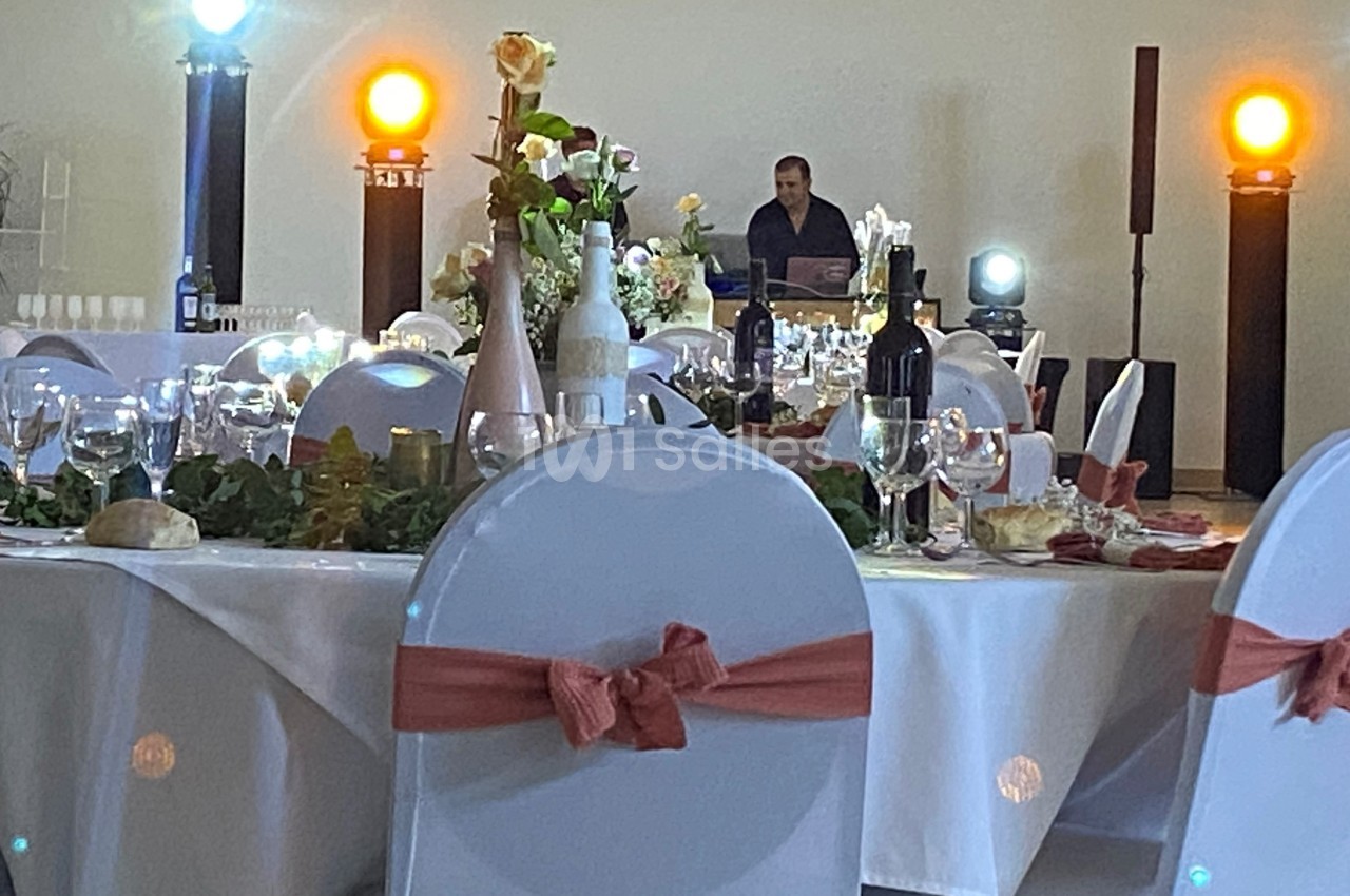 Salle de réception décorée avec des tables dressées, des chaises ornées de nœuds roses et un DJ en arrière-plan.