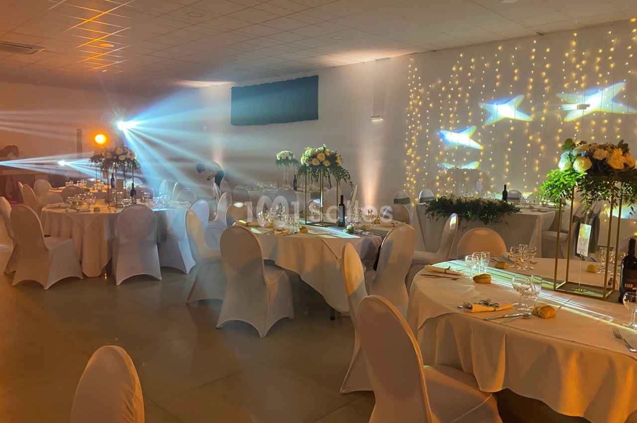 Salle de réception décorée avec des tables rondes, nappes blanches, éclairage tamisé et guirlandes lumineuses au mur.