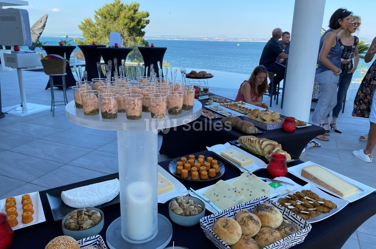 Buffet en extérieur avec vue sur la mer, comprenant pains, viennoiseries, fromages et verrines disposés sur des tables.