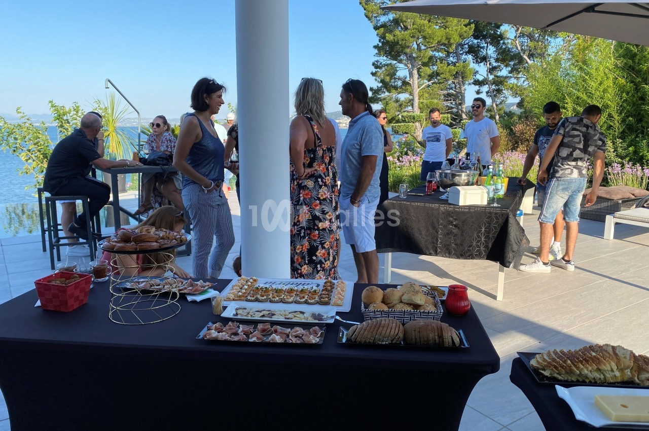 Groupe de personnes discutant autour de tables garnies de plats lors d'un événement en extérieur avec vue sur la mer.