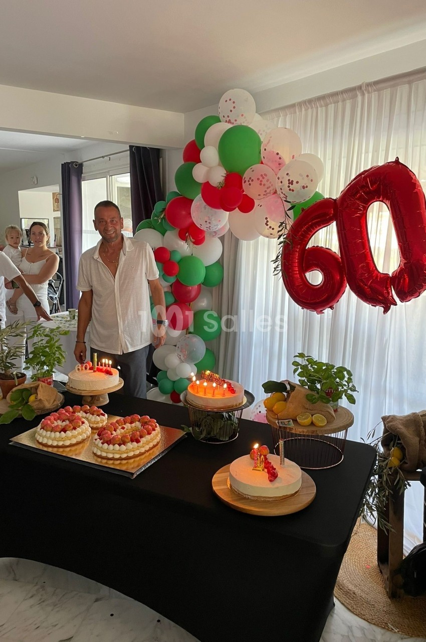 Un homme souriant devant une table décorée avec des gâteaux, des ballons et le chiffre 60 en rouge.