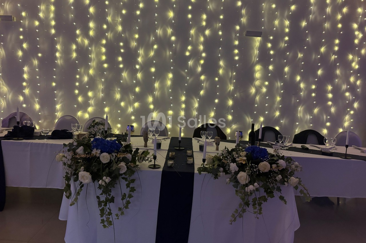 Table décorée pour un événement, ornée de fleurs blanches et bleues, avec un fond de guirlandes lumineuses.