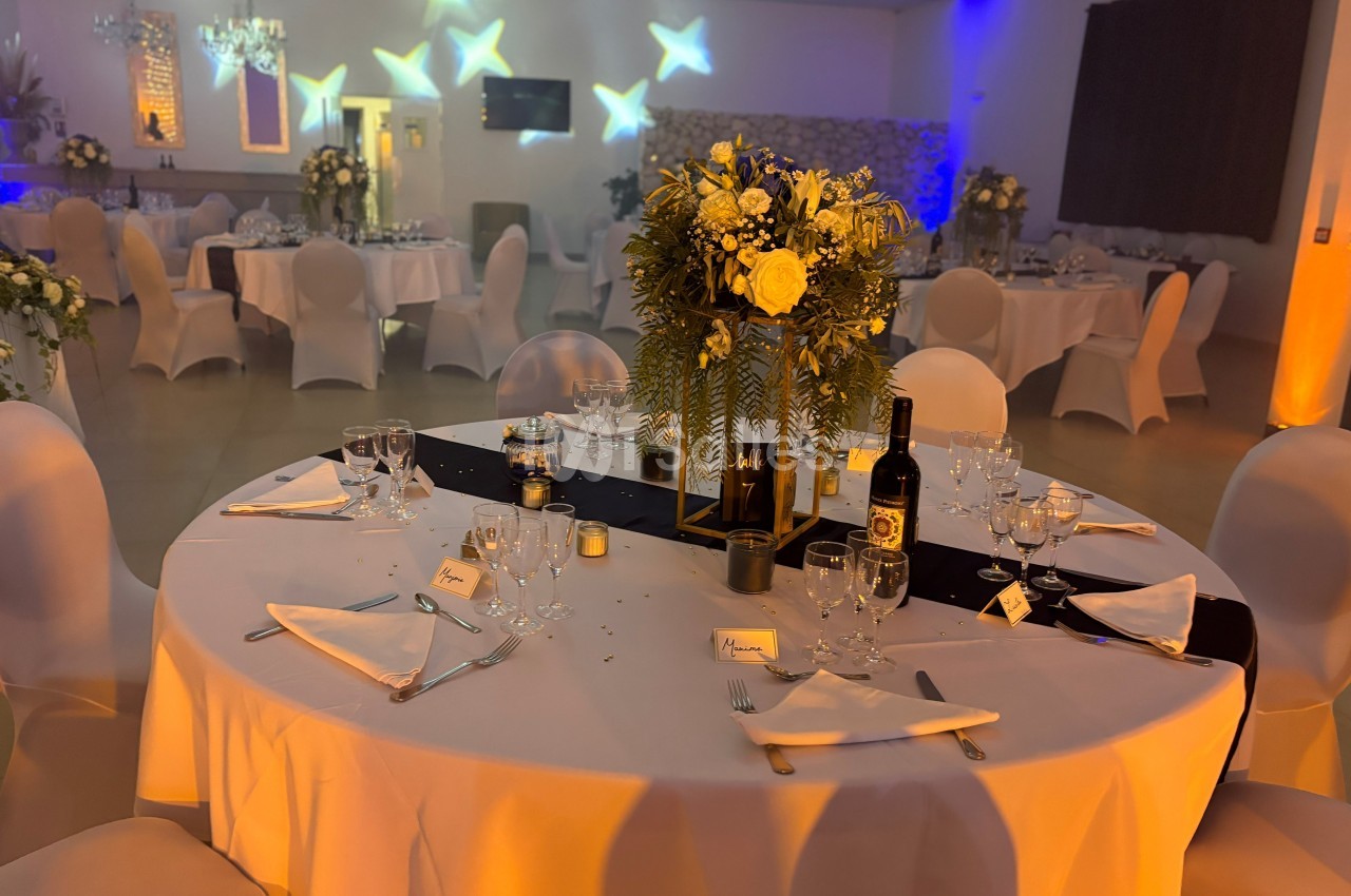 Salle de réception décorée avec des tables rondes élégantes, nappes blanches, fleurs et éclairage tamisé.