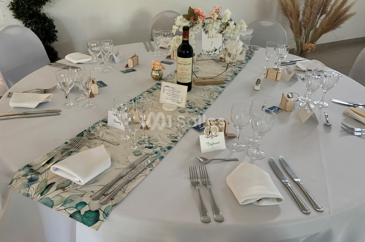 Table dressée pour un événement avec nappes blanches, décoration florale, bouteille de vin et couverts disposés.