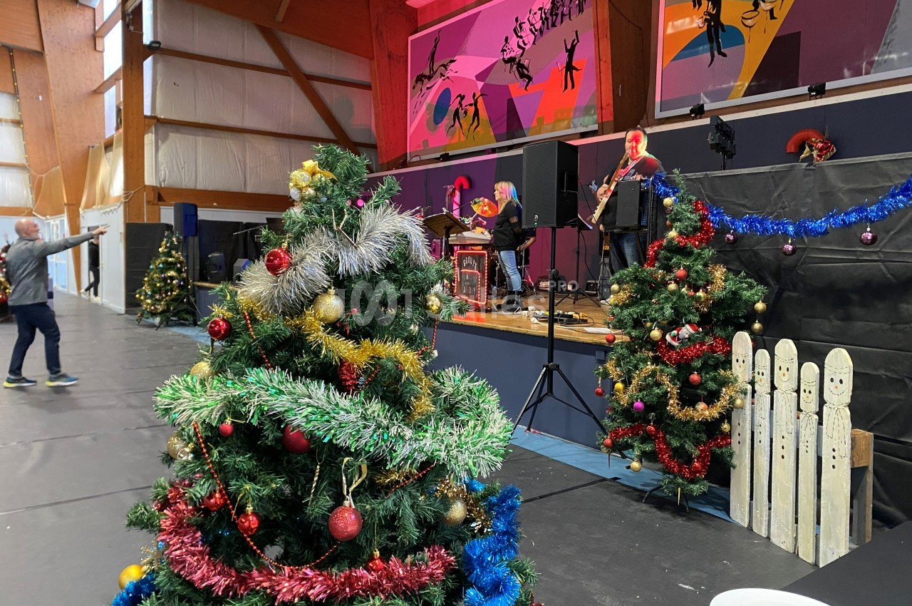 Sapin de Noël décoré au premier plan dans une salle avec scène, musiciens et décorations festives en arrière-plan.