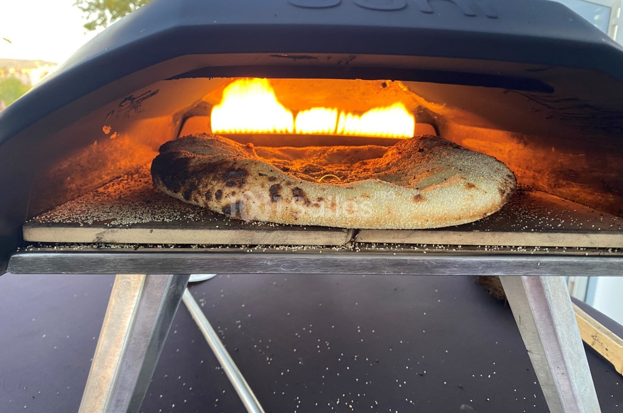 Pizza en cours de cuisson dans un four à bois, avec une flamme vive visible à l’arrière.