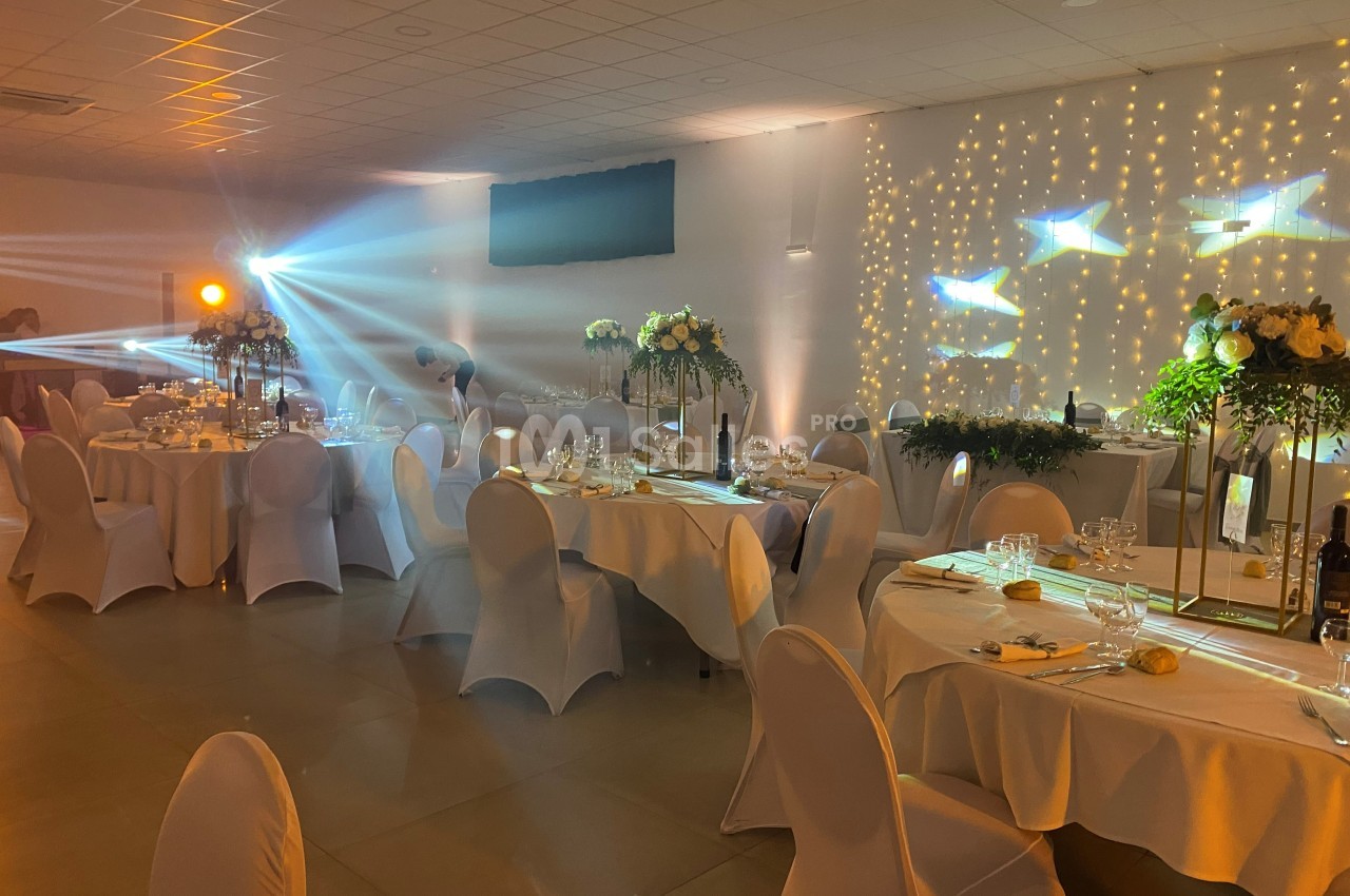 Salle de réception décorée avec des tables rondes, nappes blanches, éclairage tamisé et guirlandes lumineuses au mur.