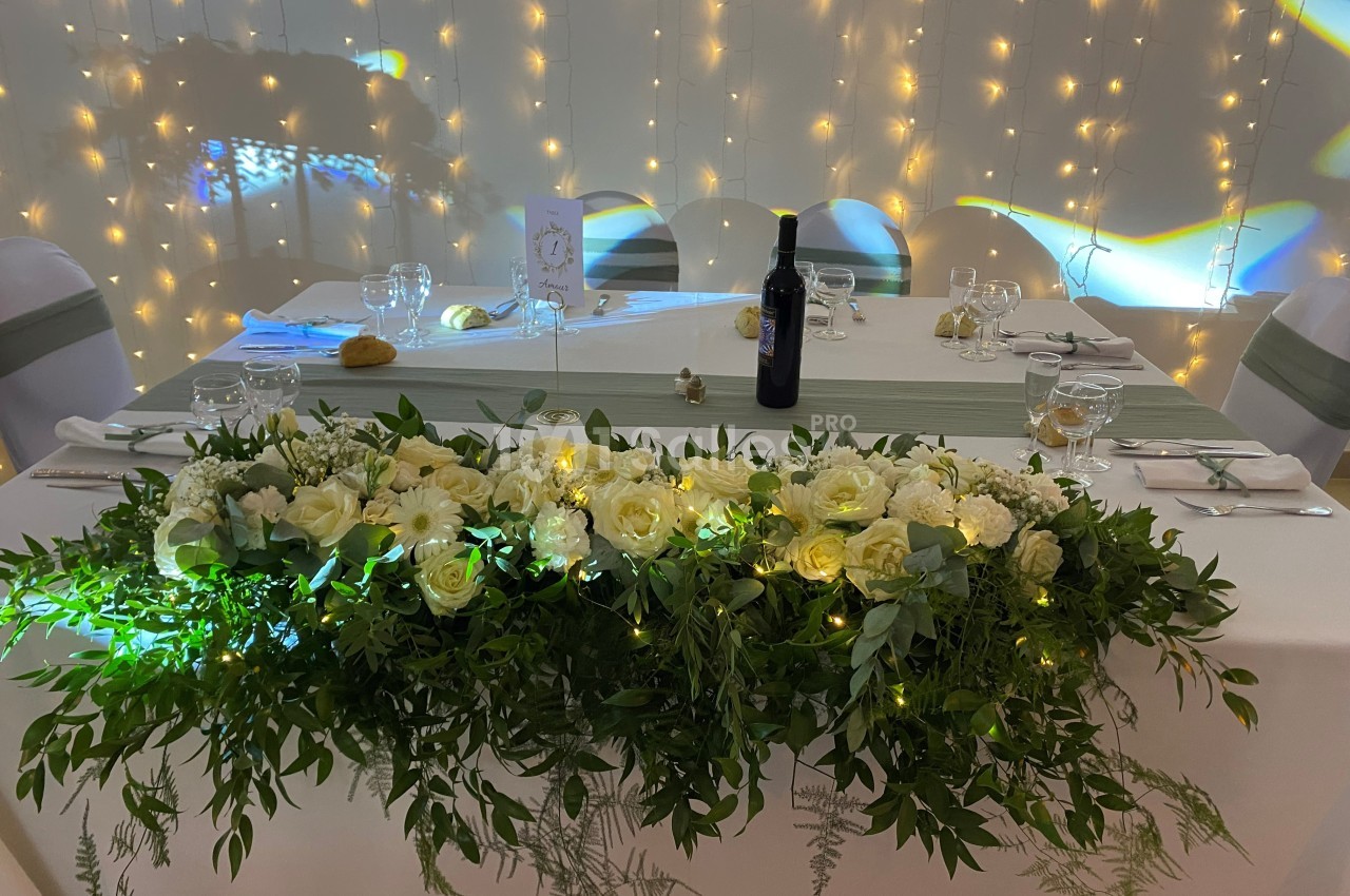 Table décorée pour un mariage avec un centre de table floral blanc et vert, éclairée par des guirlandes lumineuses.
