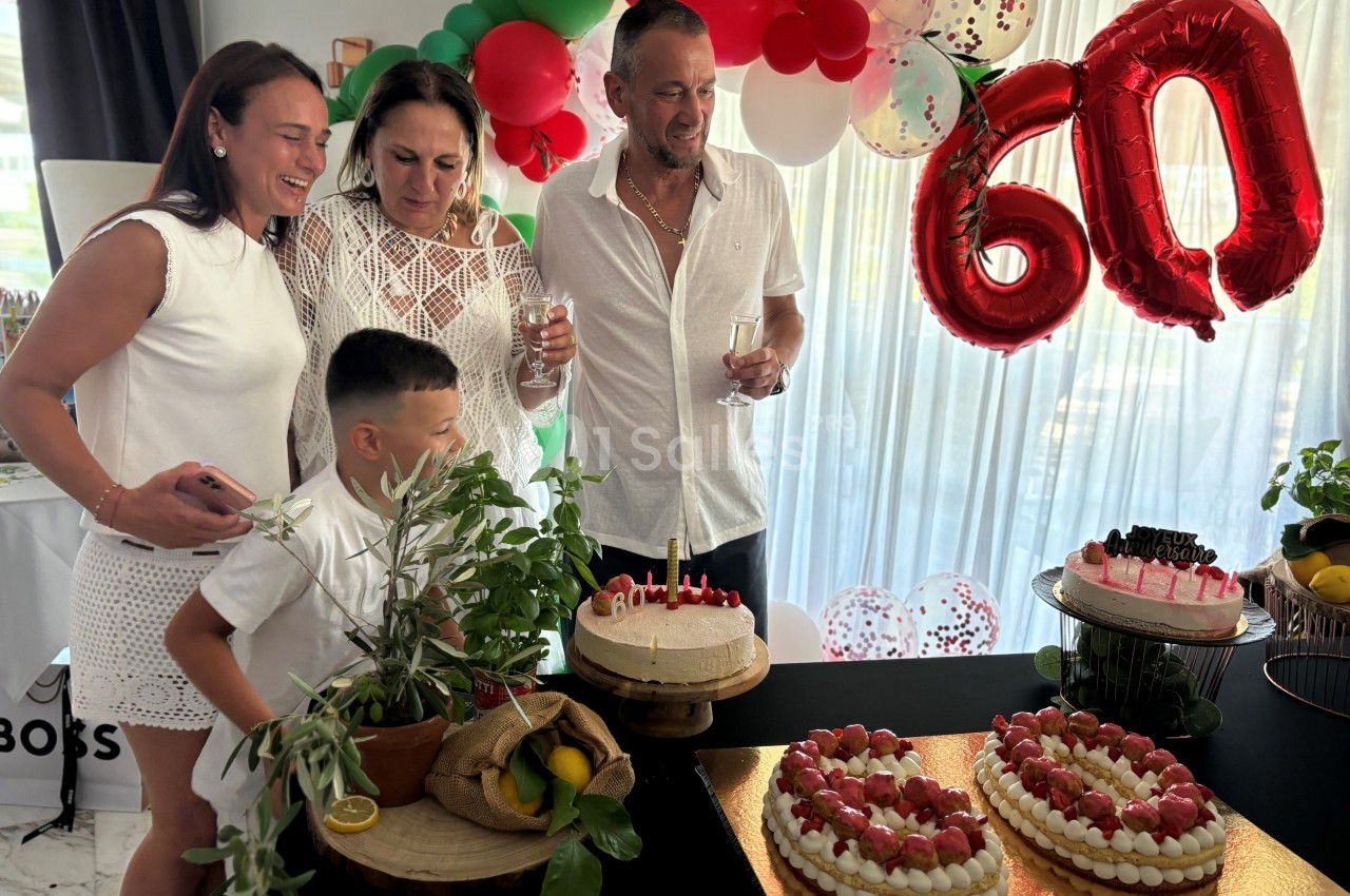 Un groupe de personnes souriantes autour de gâteaux d'anniversaire, décorations et ballons marquant ’60’ en arrière-plan.