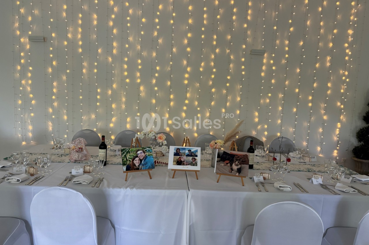 Table décorée pour un événement avec nappes blanches, guirlandes lumineuses en arrière-plan et photos sur chevalets.