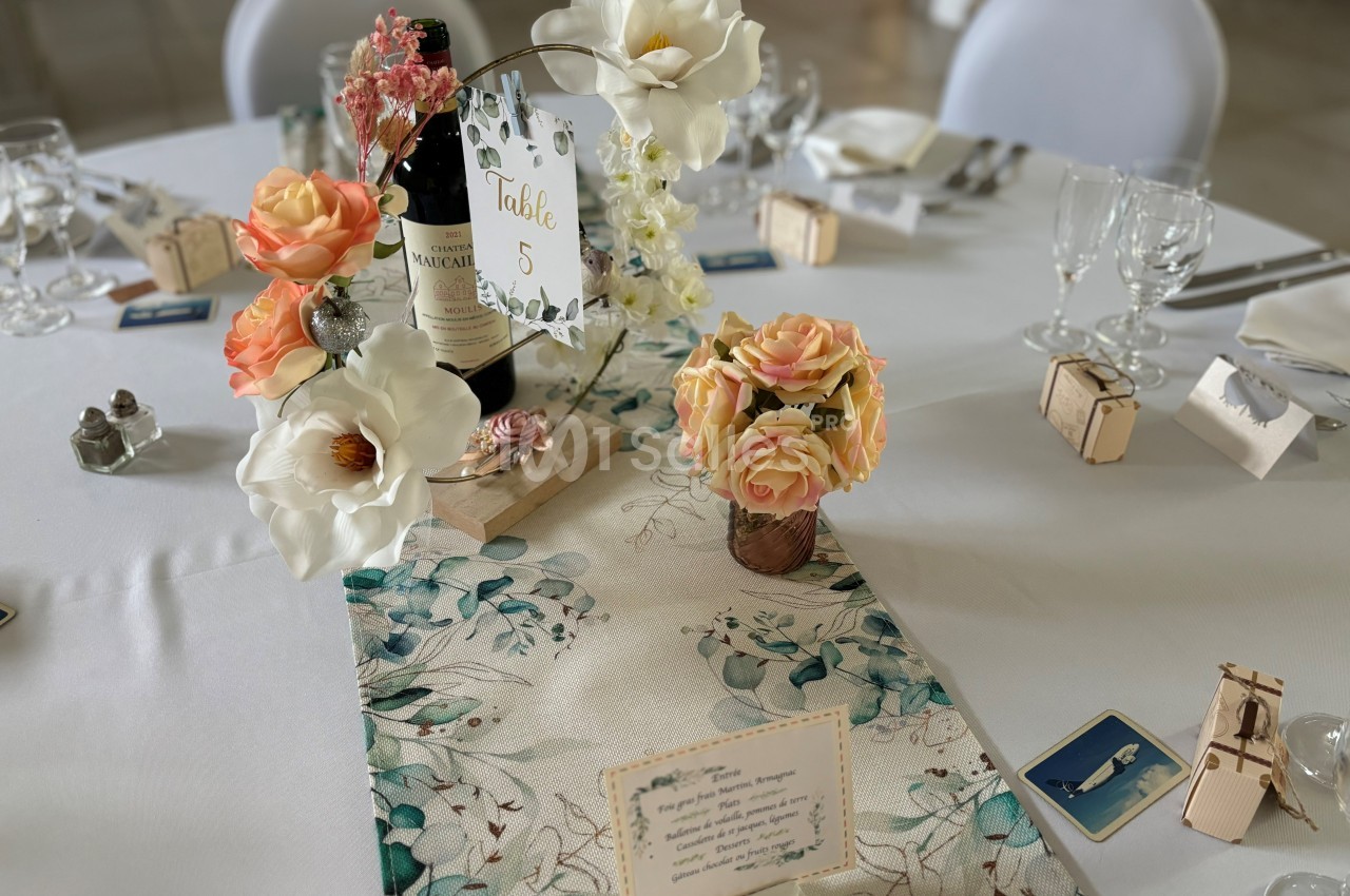 Centre de table floral avec roses et magnolias, bouteille de vin, numéro de table et décorations sur une nappe blanche.
