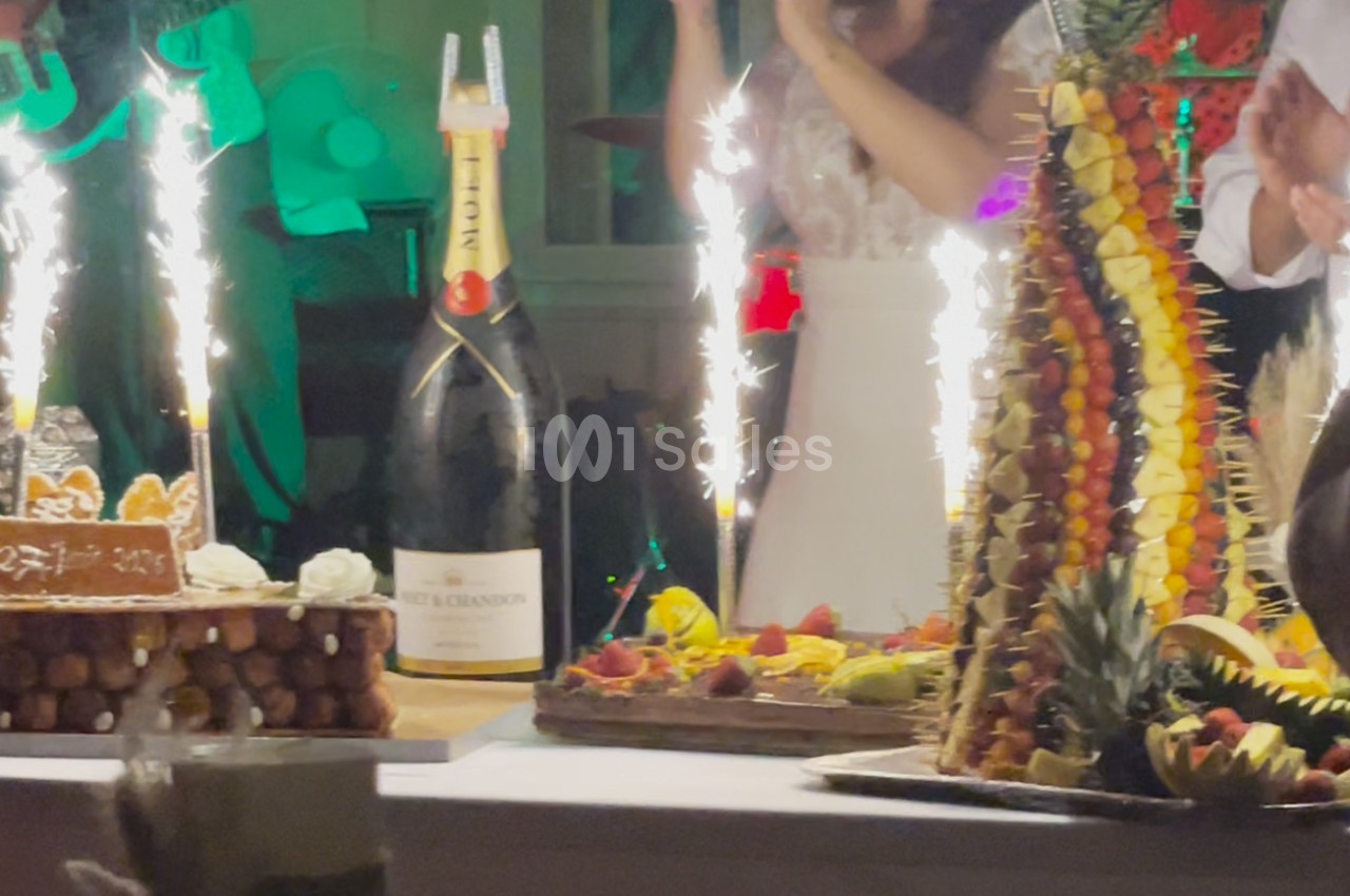 Table festive avec pièce montée de fruits, gâteau, bouteille de champagne et cierges scintillants allumés.
