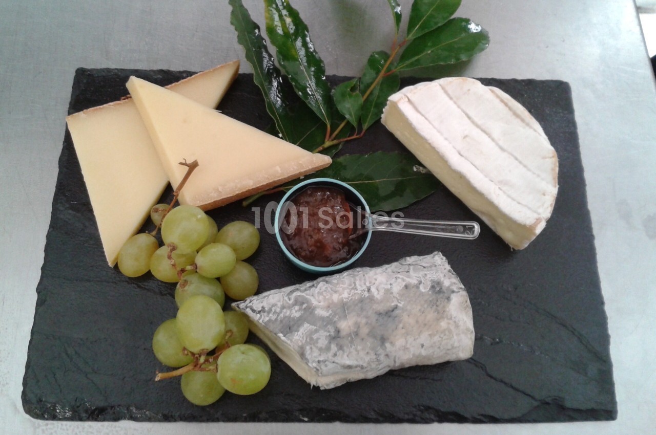 Plateau de fromages variés avec raisins, confiture et feuilles décoratives sur une ardoise noire.