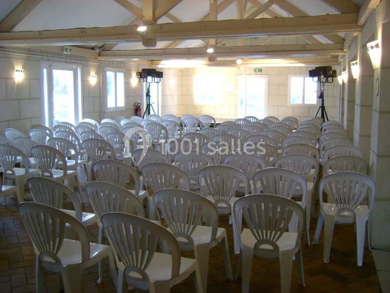 Salle avec des rangées de chaises en plastique blanc disposées face à une scène lumineuse dans un espace aux murs clairs.