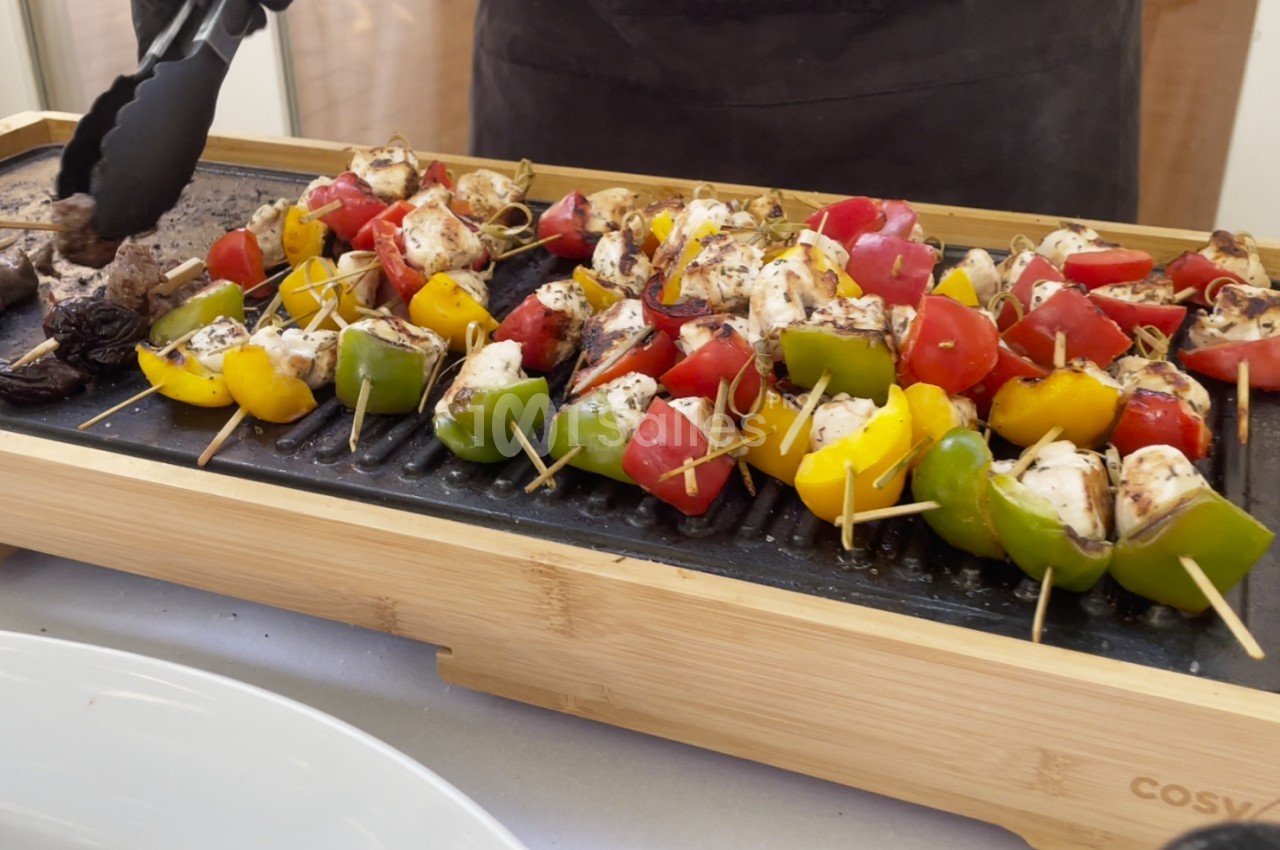 Brochettes de légumes et de poulet grillées sur une plancha en bois, avec des pinces tenant un morceau.