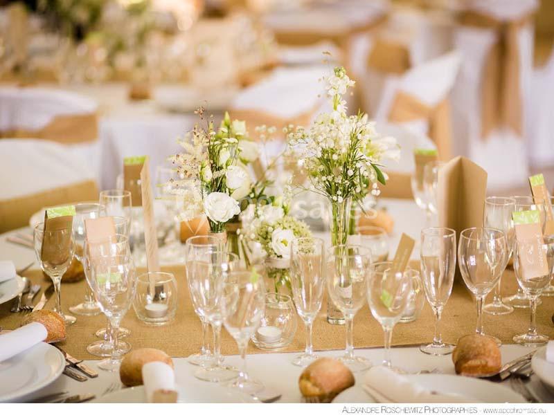 Table dressée avec nappes blanches, chemins de table en toile de jute, fleurs blanches et verres disposés pour un repas.