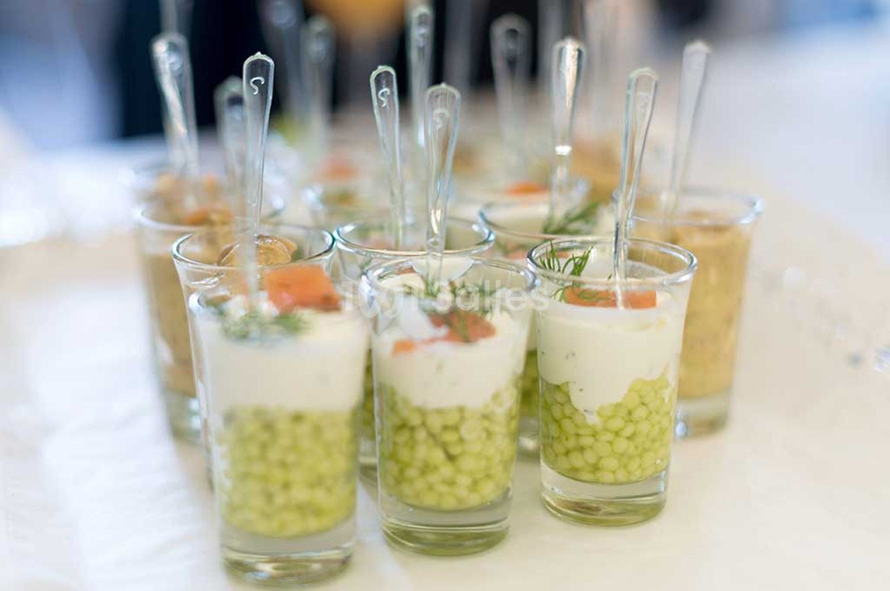 Verrines garnies de petits pois, crème, saumon fumé et aneth, présentées avec des cuillères transparentes.