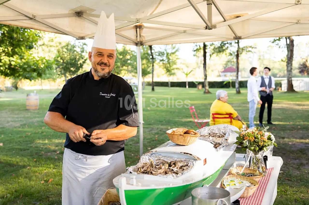 Un chef en tenue prépare des huîtres sous une tente dans un cadre extérieur verdoyant avec des invités en arrière-plan.
