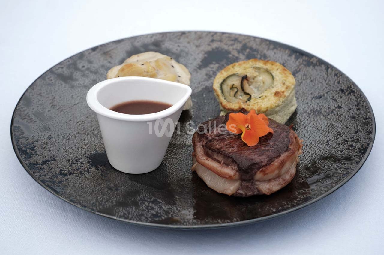 Assiette gastronomique avec viande, roulé de légumes, accompagnement et sauce servie dans une coupelle blanche.