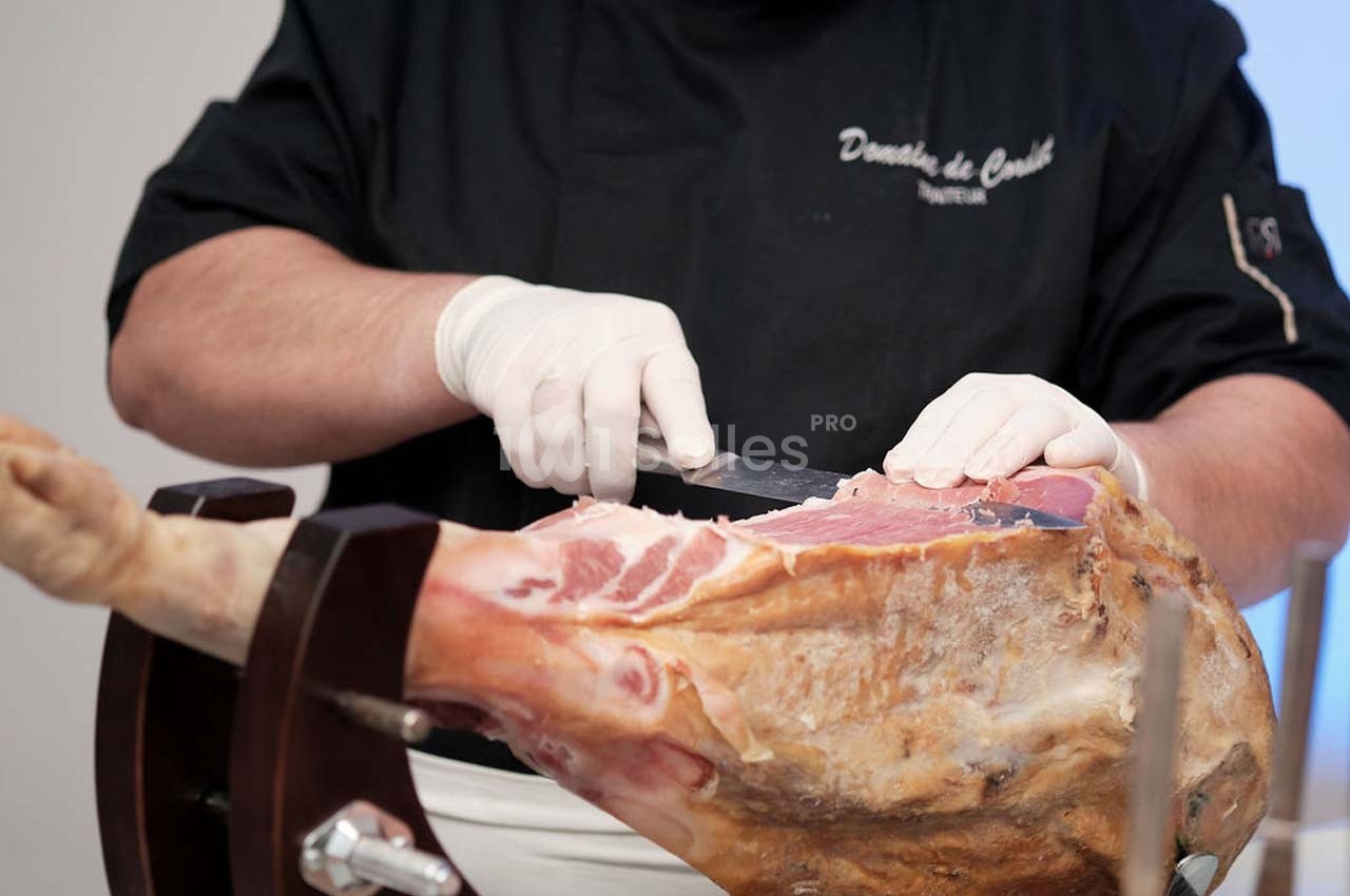 Un professionnel découpe finement un jambon cru sur un support en bois, portant des gants blancs.