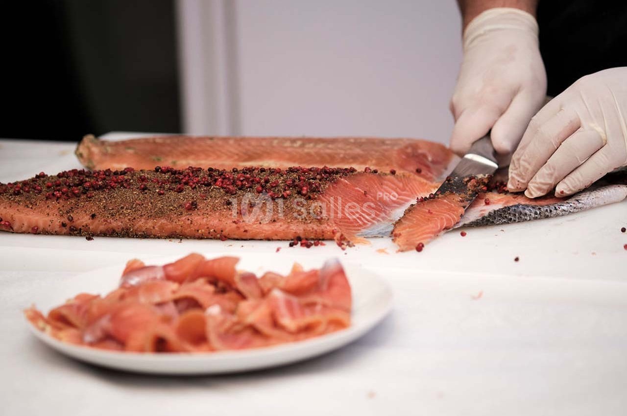 Un cuisinier tranche du saumon fumé recouvert de grains de poivre, avec des morceaux déjà servis sur une assiette.