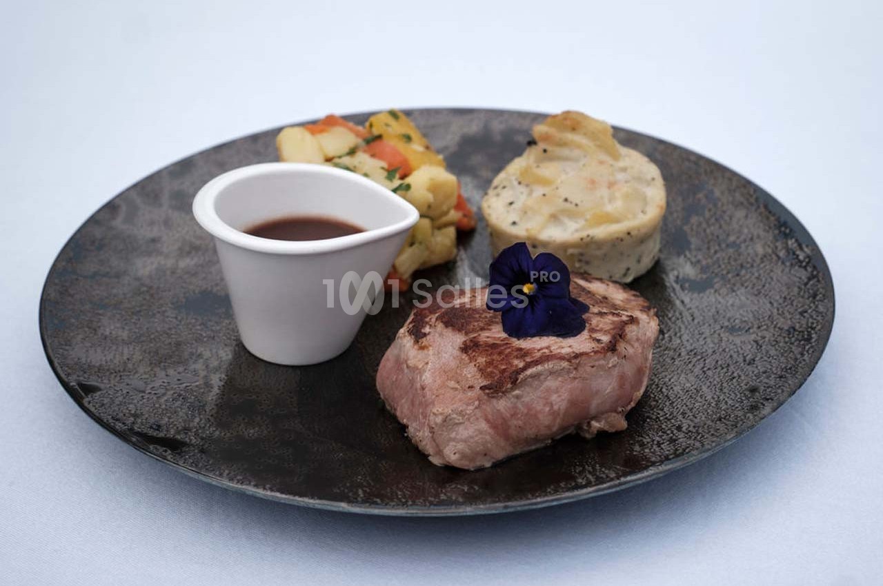 Assiette avec un filet de viande, une sauce en ramequin, des légumes et un gratin, décorée d'une fleur comestible.