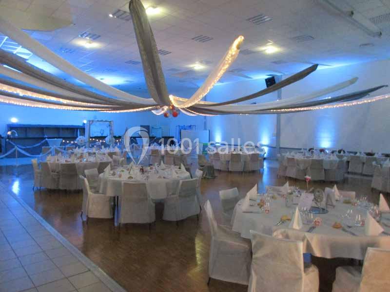 Salle de réception décorée avec des tables rondes dressées, éclairage tamisé et guirlandes lumineuses au plafond.