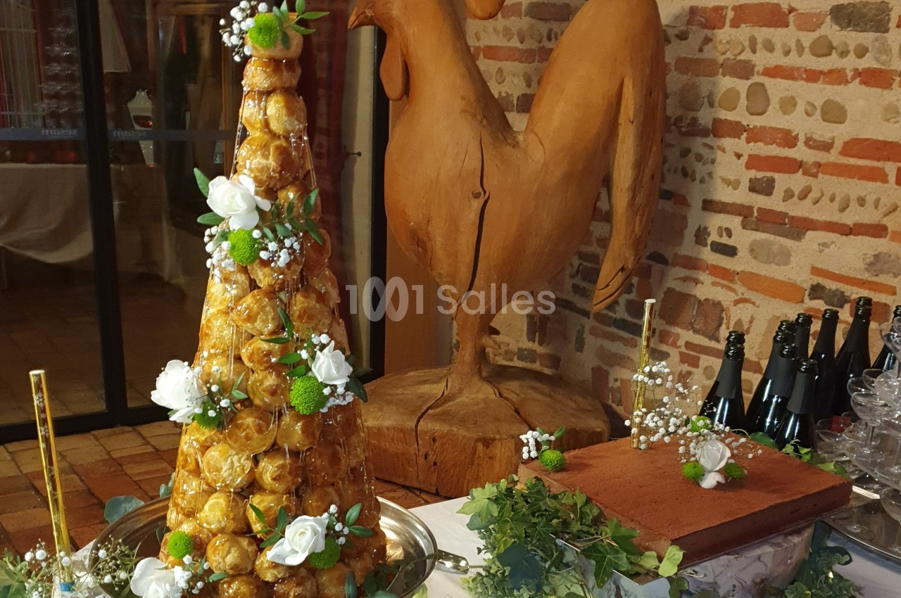 Pièce montée en choux décorée de fleurs, posée sur une table avec un coq sculpté en arrière-plan.