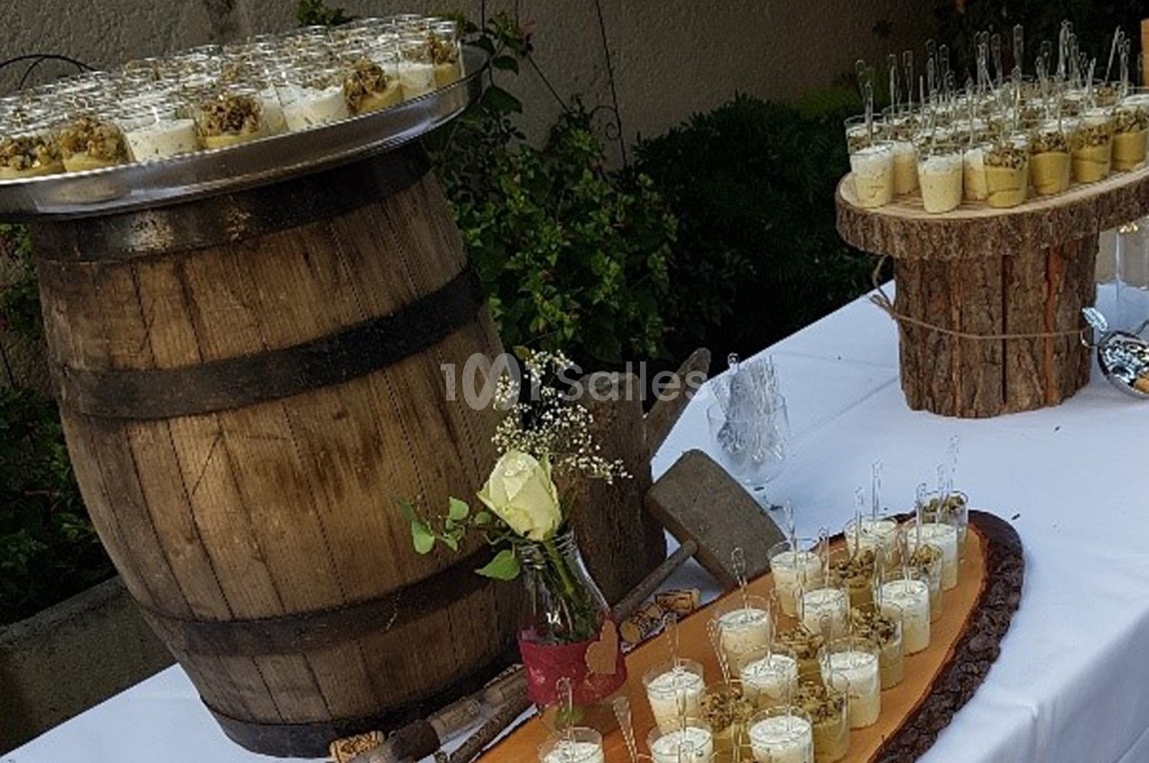 Buffet en extérieur avec verrines disposées sur une table blanche, un tonneau en bois et des rondins décoratifs.