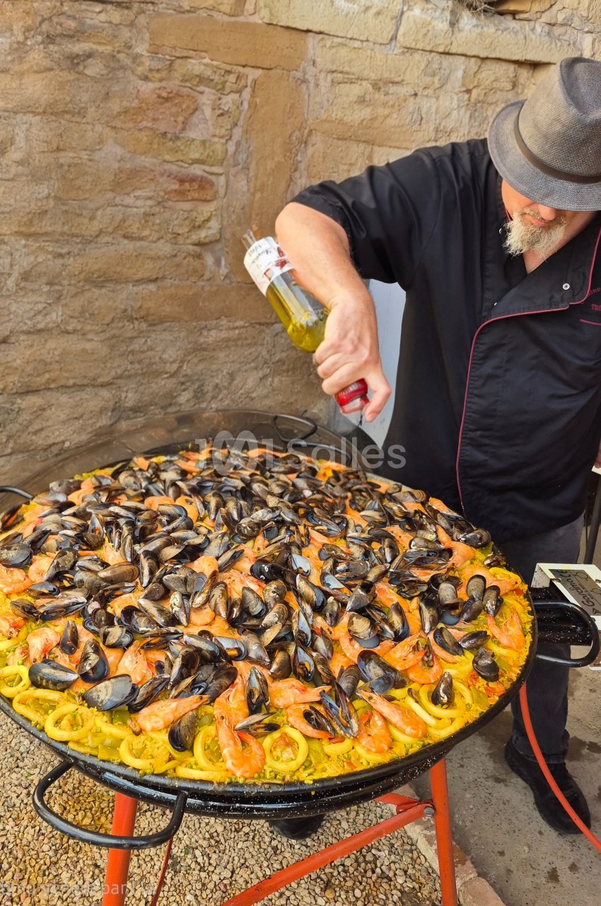 Un cuisinier prépare une grande paella garnie de moules, crevettes et calamars dans une poêle extérieure.