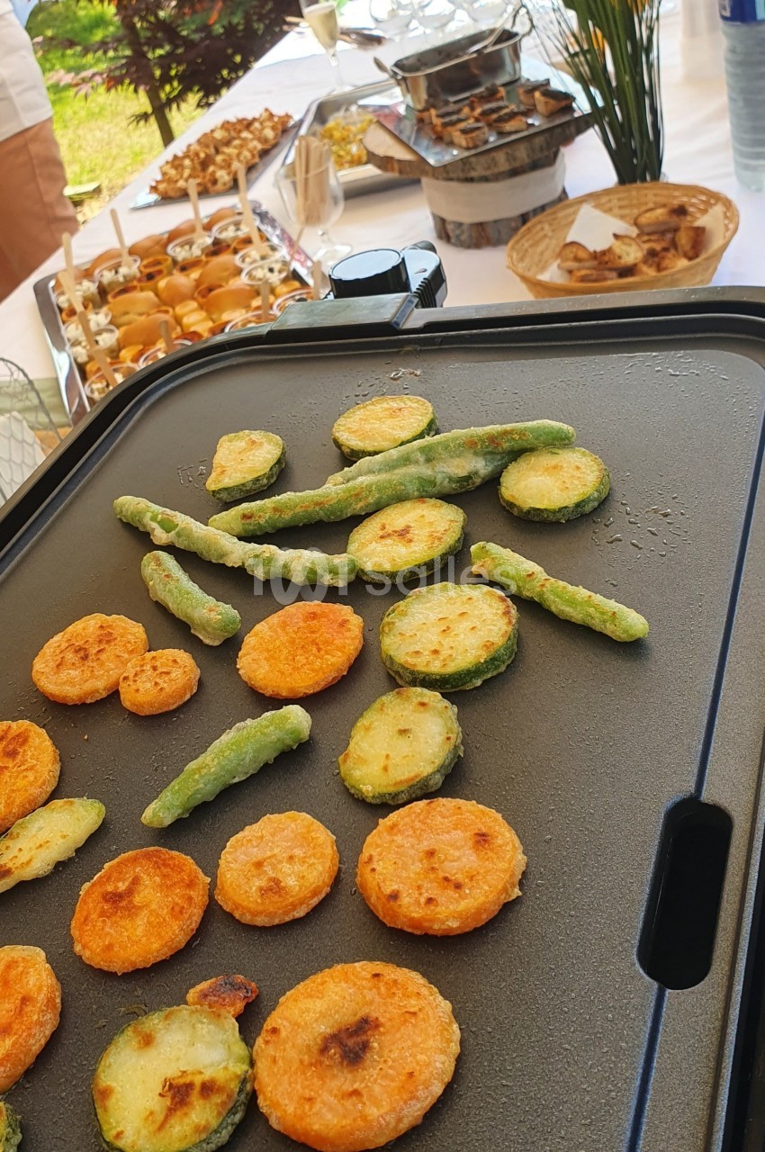 Légumes frits en cours de cuisson sur une plancha, avec un buffet varié en arrière-plan.