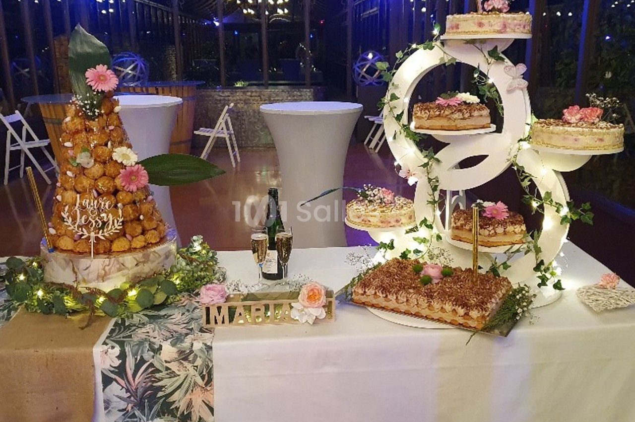 Table décorée pour un événement avec pièces montées, gâteaux, champagne et ornements floraux dans une salle éclairée.