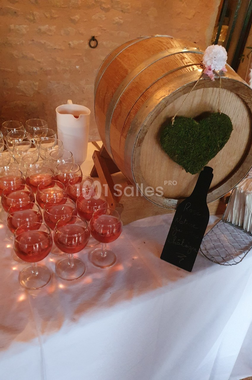 Baril en bois décoré de mousse en forme de cœur, entouré de verres de boisson rose sur une table blanche.